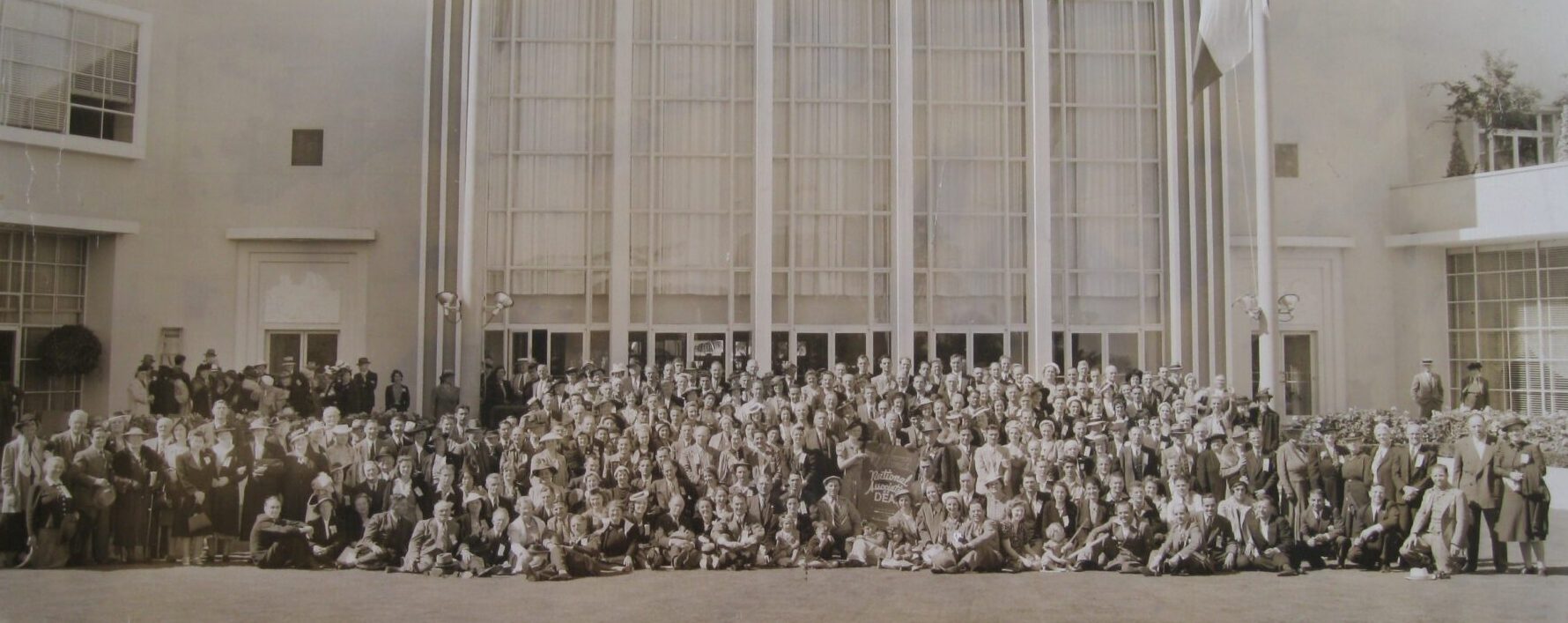 A sepia-toned vintage photograph of a large group of people gathered in front of a modernist-style building with tall windows and decorative light fixtures. Many are dressed in formal attire, with hats and suits. A small sign in the center reads &ldquo;National Association of the Deaf.&rdquo;